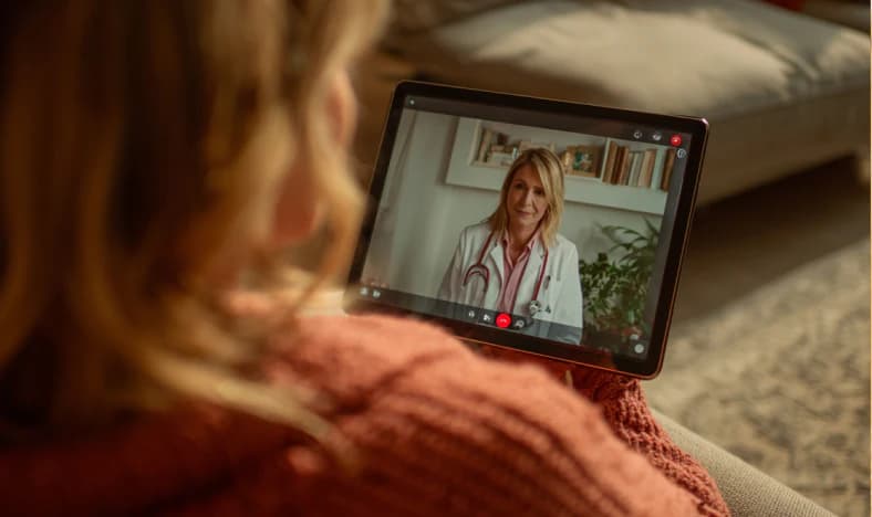 Patient on a couch holding a tablet during a telehealth video consultation with a smiling female doctor wearing a stethoscope